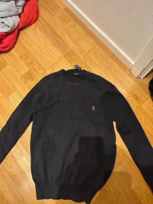 Svart tröja från Ralph Lauren - Klassisk svart tröja från Ralph Lauren med rund halsringning och den ikoniska lila broderade loggan på bröstet. Tröjan har långa ärmar och ribbade muddar vid ärmslut och nederkant. Perfekt för en stilren och enkel look. Den är lite paj vid hängaren men kan laga innan köpet!