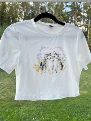 Y2K Vit baby tee med gulligt katt-tryck, stl M - En vit y2k baby tee med ett gulligt katt-tryck på framsidan 🩷 Säljer pga att den inte kommer till användning. Längd: 39cm - Axel: 35cm - Byst: 83cm