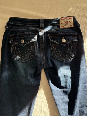 True Religion mörkblå jeans - Säljer ett par mörkblå jeans från True Religion med ikoniska kontrastsömmar och stora fickor bak med lock och silverfärgade knappar. Klassisk patch med logga bak i midjan. Snygg passform och slitstarkt denimtyg.