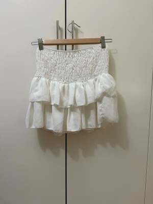 Vit volangkjol med smock - Supersöt vit kortkjol med smock upptill och dubbla volanger. Kjolen har en luftig och lätt känsla, perfekt för varma dagar. Passar till både sneakers och sandaler för en trendig look.