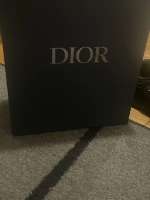 Dior b30 - Säljer en elegant mörkblå presentask från Dior med tydlig logga i silver på framsidan. Perfekt för att förvara eller ge bort exklusiva produkter. Asken har en stilren och lyxig känsla och är tillverkad i hård kartong.