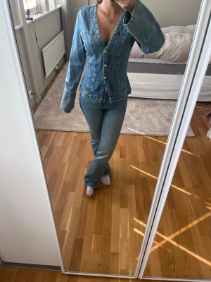 Figurnära jeansjacka med v-ringning - Snygg figurnära jeansjacka i klassisk blå denim med djup v-ringning och knappstängning framtill. Jackan har långa ärmar och markerad midja för en trendig siluett. Perfekt att styla med jeans för en cool denim-on-denim look.