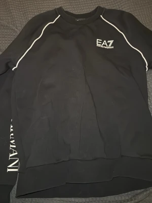 Svart sweatshirt från EA7 Emporio Armani - Snygg svart sweatshirt från EA7 Emporio Armani med vita kontrastsömmar längs axlarna och stort vitt logotryck på ena ärmen. Liten EA7-logga på bröstet. Tröjan har rund hals och är tillverkad i mjukt material, perfekt för en avslappnad streetwear-look.