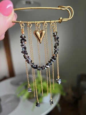 Dekorativ säkerhetsnål med kedjor - Unik säkerhetsnål i guldfärgad metall med hängande kedjor, glaspärlor och ett hjärtformat hänge. Perfekt som accessoar till jacka eller väska för en personlig touch. Kedjorna ger en bohemisk och trendig känsla.