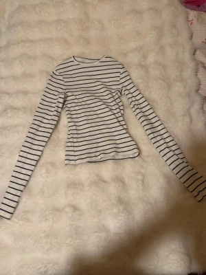 Randig långärmad topp från BikBok - Säljer en vit långärmad topp från BikBok med tunna svarta horisontella ränder. Toppen har en rund halsringning och är i ett mjukt, ribbat material som sitter snyggt och figurnära. Perfekt att styla med jeans eller kjol.