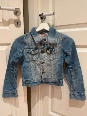 Blå klassisk barn jeansjacka, storlek 134  - Snygg blå jeansjacka med klassisk passform och knappar framtill. Jackan har två bröstfickor med lock och knapp, samt detaljerade sömmar för en cool look. Perfekt att slänga över en hoodie eller t-shirt för en avslappnad stil.
