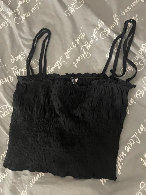 Svart smockad linnetopp Divided - Svart linnetopp från Divided by H&M med smala axelband och smockad struktur. Toppen har en lätt rynkad kant upptill och nedtill för en söt look. Perfekt att matcha med jeans eller kjol för en chill stil.