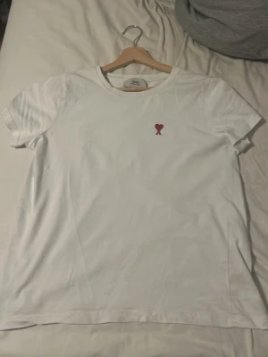 Vit t-shirt från Ami med hjärtdetalj - Säljer en vit t-shirt från Ami Alexandre Mattiussi i storlek M. T-shirten har en liten röd hjärtdetalj broderad på bröstet och klassisk rund hals. Tillverkad i mjuk bomull och har korta ärmar. Perfekt för en clean och stilren look.