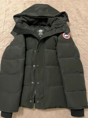 Svart dunjacka från Canada Goose - Snygg svart dunjacka från Canada Goose med avtagbar huva och klassisk logotyp på ärmen. Jackan har dragkedja och knappar framtill, samt ribbade muddar vid ärmsluten. Perfekt för kalla dagar och riktigt varm tack vare dunfyllningen. Pris kan diskuteras.