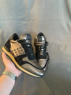 Valentino rockrunner  - Snygga sneakers från Valentino i metallic grått och svart med coola nitar på sidorna. Skorna har svarta skosnören, svart mocka och metallicläder, samt en beige sula. Perfekt för dig som vill sticka ut med ett exklusivt märke och unik design.