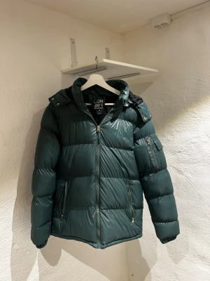 Mörkgrön pufferjacka med huva - Stilren mörkgrön pufferjacka från Brave Soul med huva och dragkedja framtill. Jackan har två framfickor med dragkedja och en extra ficka på ärmen. Tillverkad i glansigt syntetmaterial som håller värmen under kalla dagar.