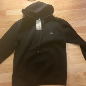 Svart hoodie från Lacoste - Snygg svart hoodie från Lacoste med klassisk krokodillogga på bröstet. Hoodien har huva med dragsko och en stor magficka framtill. Tillverkad i mjukt material som känns skönt mot huden. Perfekt för en avslappnad och stilren look.