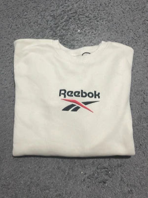 Vit sweatshirt från Reebok - Säljer en stilren vit sweatshirt från Reebok med klassisk logga i svart och rött på bröstet. Tröjan har rund halsringning och ribbade muddar vid ärmslut och nederkant. Perfekt för en avslappnad och sportig look.