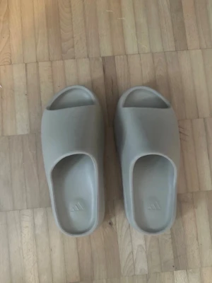 Beige slides från Adidas - Säljer ett par beigea slides från Adidas med stilren och chunky design. Tofflorna har öppen tå och är gjorda i ett mjukt, formgjutet material som ger skön komfort. Perfekta för att glida runt hemma eller till och från gymmet.