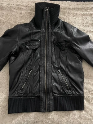 Svart skinnjacka från Bershka - Snygg svart skinnjacka från Bershka med hög krage i ribbat tyg, två bröstfickor med lock och två snedställda fickor med dragkedja. Jackan har ribbade muddar vid ärmslut och nederkant samt coola sömdetaljer framtill.