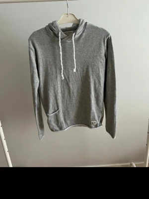 Grå hoodie med ficka och huva - En enkel och stilren grå hoodie från jack&jones med huva och vita dragsnören. Hoodien har en magficka framtill och är tillverkad i mjukt bomullsmaterial. Perfekt för en avslappnad look och passar till det mesta.
