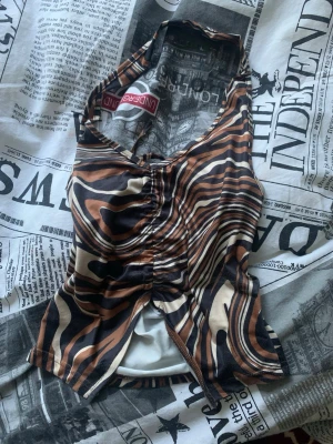 Zebramönstrad halterneck topp - Säljer en zebramönstrad halterneck topp från UNDERATED i bruna, svarta och beige toner. Toppen är ärmlös och har en croppad passform med snygg rynkning framtill. Perfekt för dig som vill sticka ut med ett djurmönster!