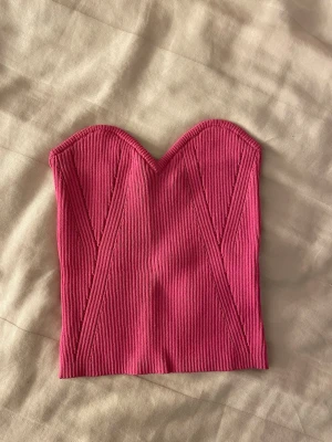 Rosa ribbad korsett-tubtopp - Trendig rosa tubtopp i korsettstil med ribbad struktur och markerade sömmar framtill. Toppen är axelbandslös och har en snygg sweetheart-ringning som ger en modern vibe. Perfekt att styla med jeans eller kjol för en cool look.