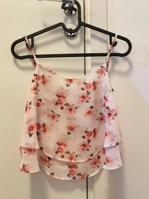 Blommig volang linnetopp - Söt linnetopp med tunna axelband och volangdetaljer. Toppen är vit med rosa blommigt mönster och har en luftig, lätt känsla. Perfekt för varma dagar och ger en romantisk vibe till din outfit.