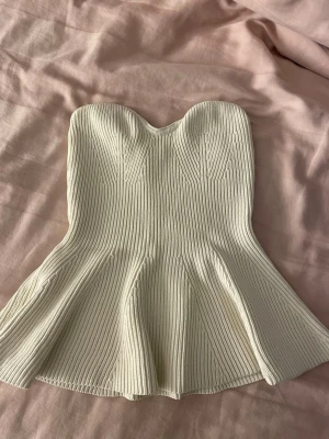 Vit ribbad axelbandslös topp - Supersnygg vit axelbandslös topp med ribbad struktur och utsvängd nederdel. Toppen har en korsettliknande form som framhäver midjan och ger en trendig siluett. Perfekt att styla med jeans eller kjol för en cool look.