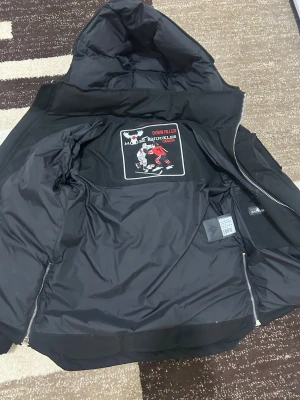 Svart dunjacka från Moose Knuckles - Säljer en svart dunjacka från Moose Knuckles med huva och dragkedja. Jackan har en klassisk design med diskreta logodetaljer på insidan och är fylld med dun för extra värme. Perfekt för kalla vinterdagar och riktigt snygg till streetwear-stilen.