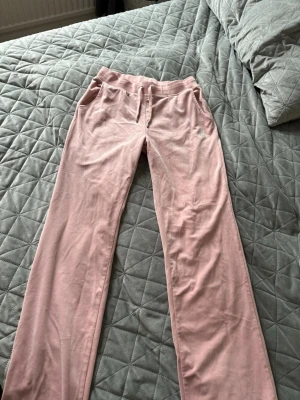 Rosa Juicy Couture mjukisbyxor - Säljer ett par klassiska rosa mjukisbyxor från Juicy Couture i mjuk velour. Byxorna har raka ben, dragsko i midjan och fickor fram. På ena fickan finns Juicy Couture-loggan i vita stenar för extra bling. Perfekta för en chill och trendig look.