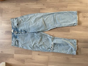 Ljusa mom jeans från H&M - Säljer ett par ljusblå mom jeans från H&M med hög midja och raka ben. Jeansen har slitningar vid ena knät och klassiska fem fickor. Tillverkade i denim med råa kanter vid benslut. Perfekta för en avslappnad och trendig look.