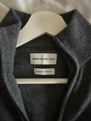 Grå merinoullströja These Glory Days - Mörkgrå ziptröja från These Glory Days i Superior Merino-ull. Tröjan har hög krage, ribbade muddar och hel dragkedja framtill. Perfekt för lager på lager och riktigt skön tack vare det mjuka materialet.