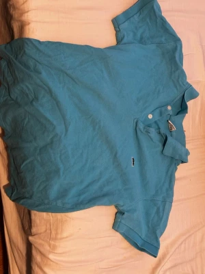 Blå pikétröja från Lacoste - Snygg blå pikétröja från Lacoste med klassisk krage och två knappar framtill. Tröjan har den ikoniska lilla krokodilloggan broderad på bröstet. Tillverkad i mjuk bomull som känns skön mot huden. Perfekt till jeans eller shorts för en avslappnad look.