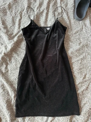 Svart kort klänning från H&M Divided - Säljer en svart kort klänning från H&M Divided med tunna axelband och v-ringning. Klänningen har en enkel, slim passform och är tillverkad i ett mjukt, glittrigt material som ger en snygg look.