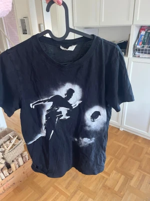 Svart t-shirt med fotbollstryck - Cool svart t-shirt med vit tryck av en fotbollsspelare som sparkar en boll. Klassisk rund hals och korta ärmar. Perfekt för dig som gillar sportig stil och vill sticka ut med ett unikt motiv.