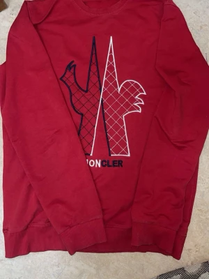 Röd hoodie från Moncler med logga - Snygg röd hoodie från Moncler med stor broderad logga på ryggen i vitt och svart. Hoodien har en klassisk passform och är tillverkad i mjukt material som känns skönt mot huden. Perfekt för dig som vill ha en statement-piece i garderoben.Storlek M