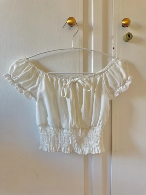 Vit offshoulder croppad topp - Söt vit offshoulder topp med korta puffärmar och smockad midja. Toppen har en liten knytning framtill och volangdetaljer vid ärmslut och nederkant. Perfekt för sommarens alla tillfällen och ger en romantisk vibe.