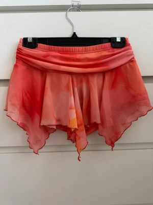 Rosa-orange kjol med asymmetrisk fåll - Säljer en unik kjol i tunnt, flowy material med rosa och orange toner. Shorts inbyggda under. Modellen har en omlott-look med asymmetrisk, vågig fåll och bred resår i midjan. Perfekt för dig som gillar färg och statement-plagg! 🧡🌺🐬