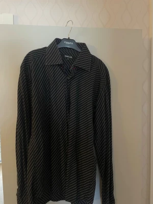 Svart randig skjorta från Emilio - Snygg svart skjorta från Emilio med tunna vita ränder och klassisk krage. Skjortan har långa ärmar och knäppning framtill. Perfekt för dig som gillar stilrena och tidlösa plagg. Skjortan är i storlek XL men passar även L. 