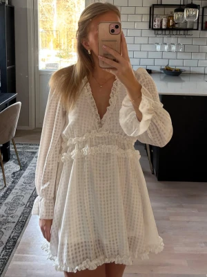 Vit kortklänning med volang  - Superfin vit kortklänning med diskret rutmönster, v-ringad hals och volangdetaljer längs ringning, midja och fåll. Långa ärmar med lätt puff och volang vid ärmslut. Perfekt till student, skolavslutning och sommarens alla tillfällen! Klänningen är använd en gång och är i mycket bra skick. Stretchig i midjan. Jag är 163 cm lång. Publicerad på flera säljsidor. 