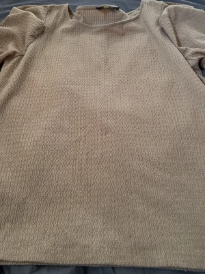 Beige topp med strukturmönster - Säljer en beige topp med vågigt strukturmönster och rund hals. Toppen har puffiga trekvartsärmar och är gjord i ett mjukt material som känns skönt mot huden. Perfekt för dig som gillar neutrala färger och en enkel men snygg look.