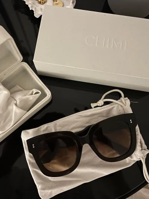 CHIMI 08 - Stilrena bruna/svarta solglasögon från CHIMI med bred båge och fyrkantig form. Glasen är mörka och bågarna har små silvriga detaljer vid fronten. Kommer med originalfodral och tygpåse för förvaring.
