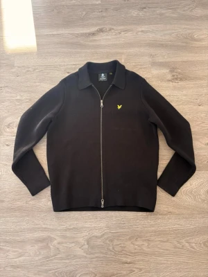 Svart ziptröja från Lyle & Scott - Snygg svart ziptröja från Lyle & Scott med gul logga på bröstet. Tröjan har hel dragkedja, klassisk krage och långa ärmar. Perfekt till lager på lager och enkel att matcha med olika outfits.