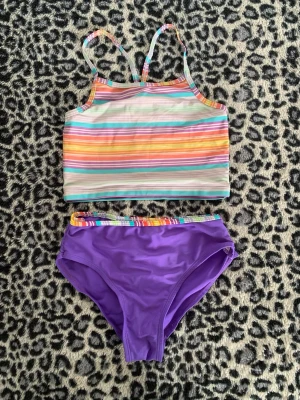 Randig bikini med lila underdel - Färgglad bikini med randigt linne i orange, rosa, blått och grönt samt lila trosa. Toppen har smala axelband och hög hals, trosan har matchande randig kant upptill. Perfekt för strandhäng eller poolen!