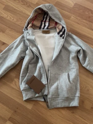 Burberry grå zip hoodie - Snygg grå zip hoodie från Burberry med klassiskt rutigt foder i huvan. Hoodien har dragkedja framtill, två fickor och ribbade muddar. Tillverkad i mjukt bomullsmaterial och har en exklusiv känsla med märkets ikoniska detaljer i huvan.