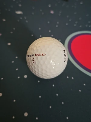Wilson Deep Red golfboll - En vit golfboll från Wilson med texten 'DEEP RED' och siffran 5 i rött. Klassisk dimple-design för bra flyg och känsla. Perfekt för dig som gillar golf och vill ha en boll med tydlig märkning.