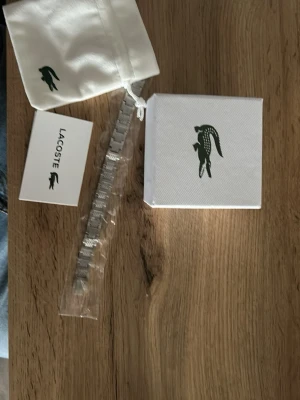 Silverfärgad länkarmband från Lacoste - Snyggt silverfärgat länkarmband från Lacoste, perfekt som reservdel eller för att byta ut ditt nuvarande armband på din Lacoste-klocka. Tillverkad i metall med klassisk design och levereras med originalpåse och ask.