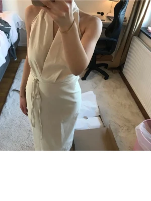 Beige omlottklänning med knyt - Säljer en snygg beige omlottklänning med halterneck och knyt i midjan. Klänningen har en elegant, lång siluett och är ärmlös, perfekt för sommarens alla tillfällen. Materialet känns mjukt och lätt mot huden.