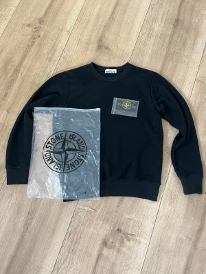 Svart sweatshirt från Stone Island - Svart långärmad sweatshirt från Stone Island med klassisk rund hals och ribbade muddar. Tröjan har den ikoniska avtagbara Stone Island-loggan på vänster ärm, fästs med knappar. Materialet är mjuk bomull, perfekt för chill dagar. 