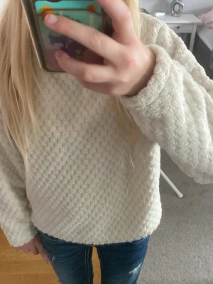 Vit fluffig stickad tröja - Mysig vit stickad tröja med fluffig struktur och hög krage. Tröjan har lång ärm och ett diskret mönster i materialet som ger en extra mjuk känsla. Perfekt för kyliga dagar och enkel att matcha med jeans eller kjol.
