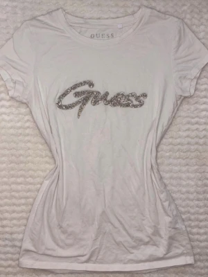 Guess t-shirt – vit med glittrig logo (S) - Vit t-shirt från Guess med snygg glittrig logga framtill. Stilren och lättmatchad – passar perfekt till både vardag och fest ✨  Storlek S, skön passform och i fint skick. Säljs billigt för snabb affär! 💨