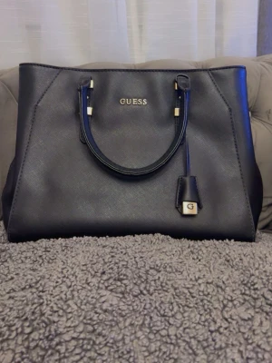 Svart handväska från Guess - Snygg svart handväska från Guess med guldfärgade detaljer och dubbla handtag. Väskan har ett rymligt innerfack med ljusrosa foder och dragkedja. Klassisk och stilren design med Guess-logga framtill och dekorativ hängare.