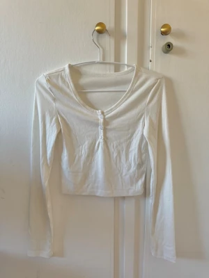 Vit croppad långärmad topp - Säljer en enkel och stilren vit croppad topp med lång ärm och rund halsringning. Toppen har tre dekorativa knappar framtill och är gjord i mjukt bomullsmaterial. Perfekt att matcha med höga jeans eller kjol.