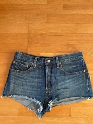 Levi's 501 blå jeansshorts med fransar i storlek W30 - Klassiska blå jeansshorts från Levi's 501 med råa fransiga kanter och femficksmodell. Storlek W30. Shortsen har normal passform och är tillverkade i slitstark bomull. Perfekta för sommaren och har den ikoniska Levi's-lappen bak.
